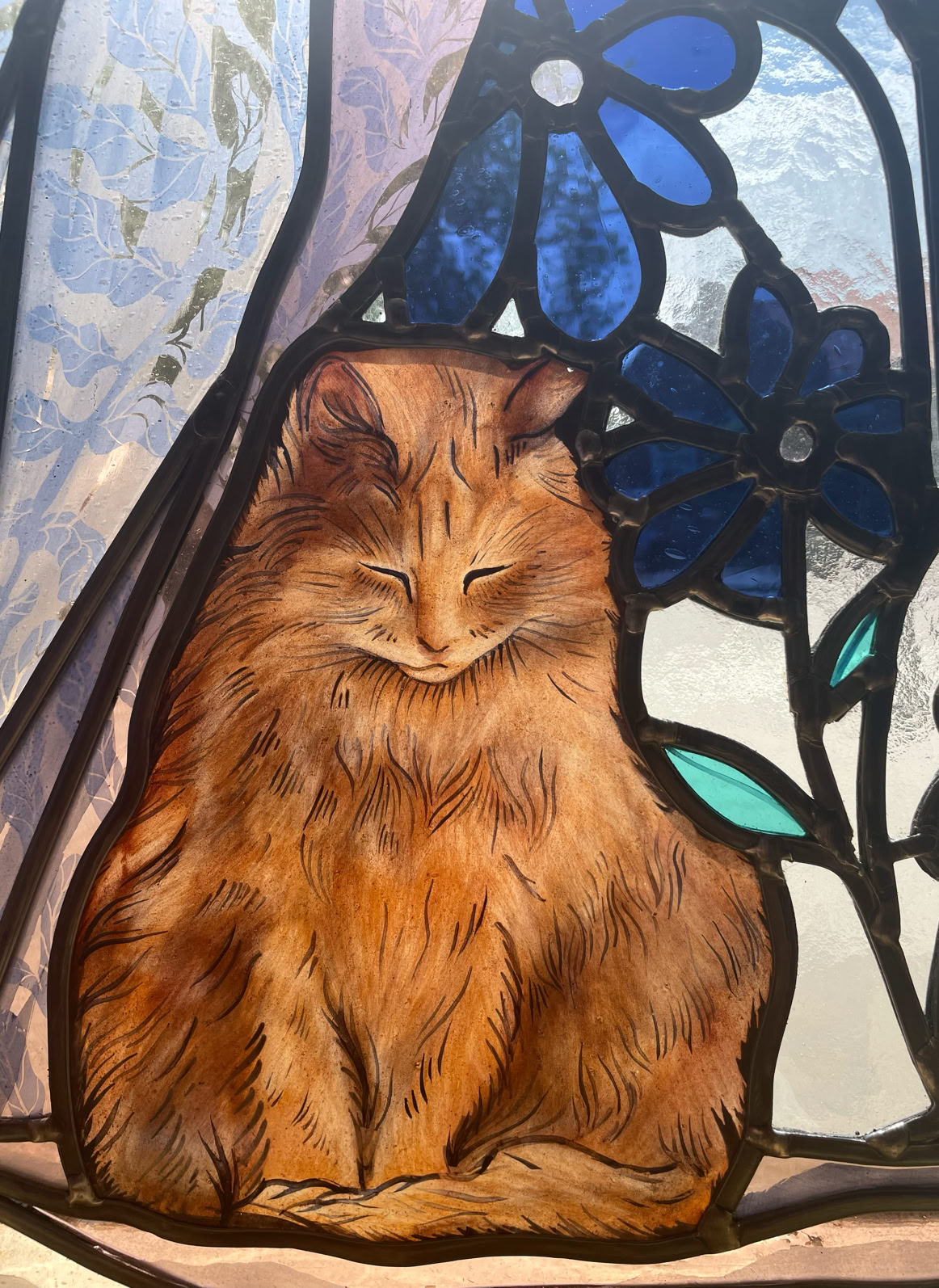 Stained glass artwork depicting a sitting orange cat with closed eyes, surrounded by blue flowers and abstract shapes.